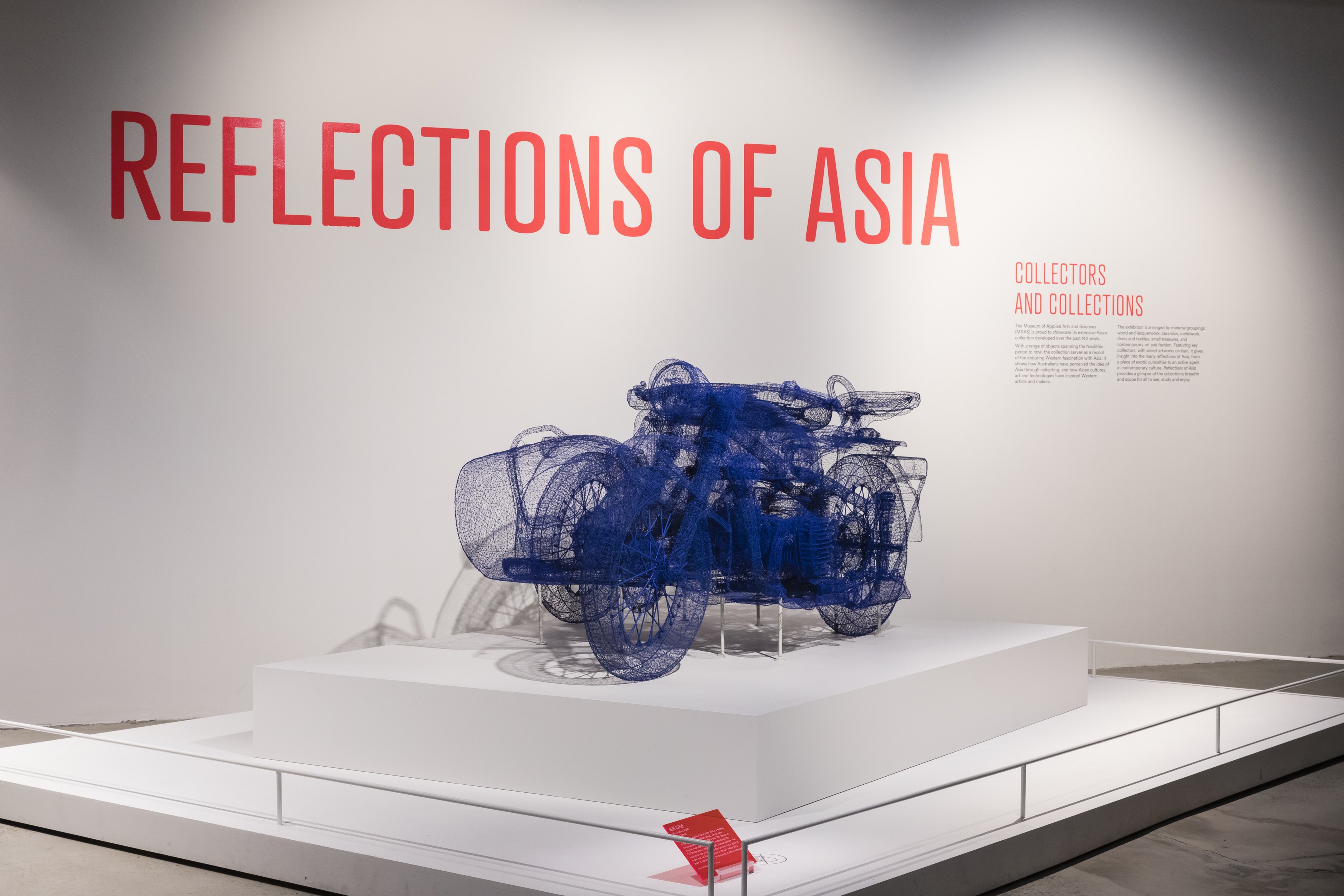 View of an exhibition title ‘Reflections of Asia: Collectors and Collection' on a white wall and an art work that is a motorcycle made of blue wire.